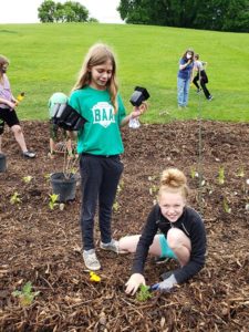 Image of two girls planting flowers in a mulched garden bed. One is holding empty pots, and one is crouched down, smiling at the camera