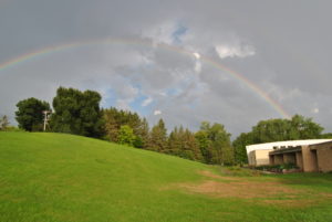A large grassy hill slopes from the left to the right, where a raingarden is planted by a school building. A double rainbow arcs overhead, seeming to land on the school.
