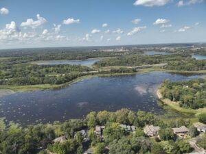 drone image of lake. clear blue water with some floating aquatic vegetation on the edges. Houses and trees on the shoreline