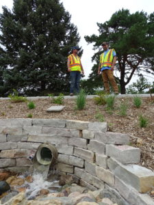 Water pours out of a spout while two people in high visibility vests stand on the hill above it.