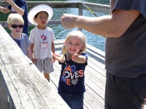 Image of three children on a fishing pier. The child in front holds out her hands for a fish on a line.