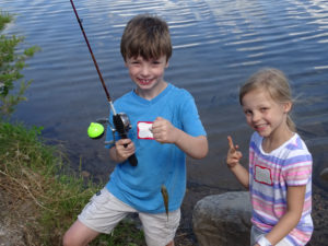 Image of two children standing by a lake. One holds a fishing rod, the other is giving the thumbs up sign.