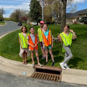 A group of girls wearing safety vest stand in front of a stormdrain
