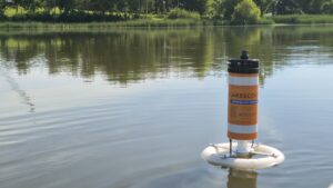 a picture of calm water with the tree shoreline reflected in the background and a black, white and orange buoy floating in the foreground.
