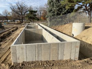 Image of a concrete structure in the midst of bare dirt with trees surrounding it. Structure has three chambers on the left, and six on the right.