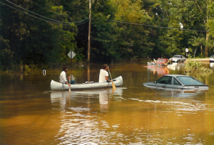 two people canoeing past a car poking out of flood waters in the street,